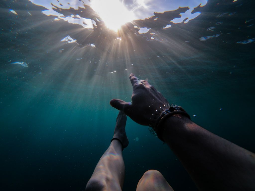 Picture taken from below the surface of the ocean, sun rays poking through, from the point of view of a person who is drowning, reaching out to the light. One arm and leg are visible, outstretched. 