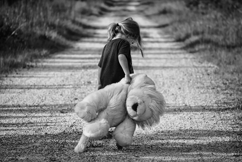 Picture of a small girl, head down, dragging an oversized teddy bear along a gravel path